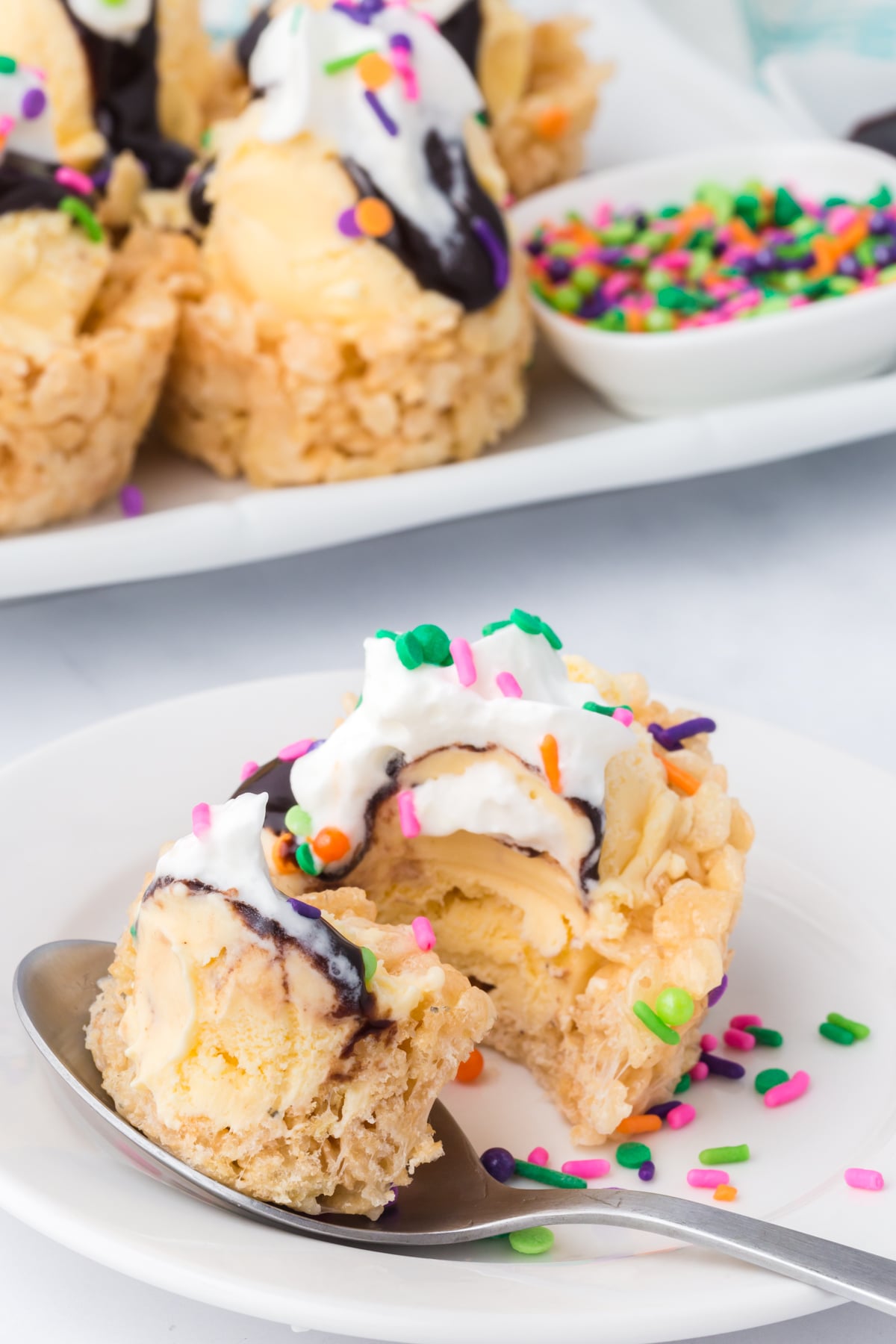Spoonful of ice cream bowl on plate with vanilla ice cream and chocolate topping in front of a platter full.