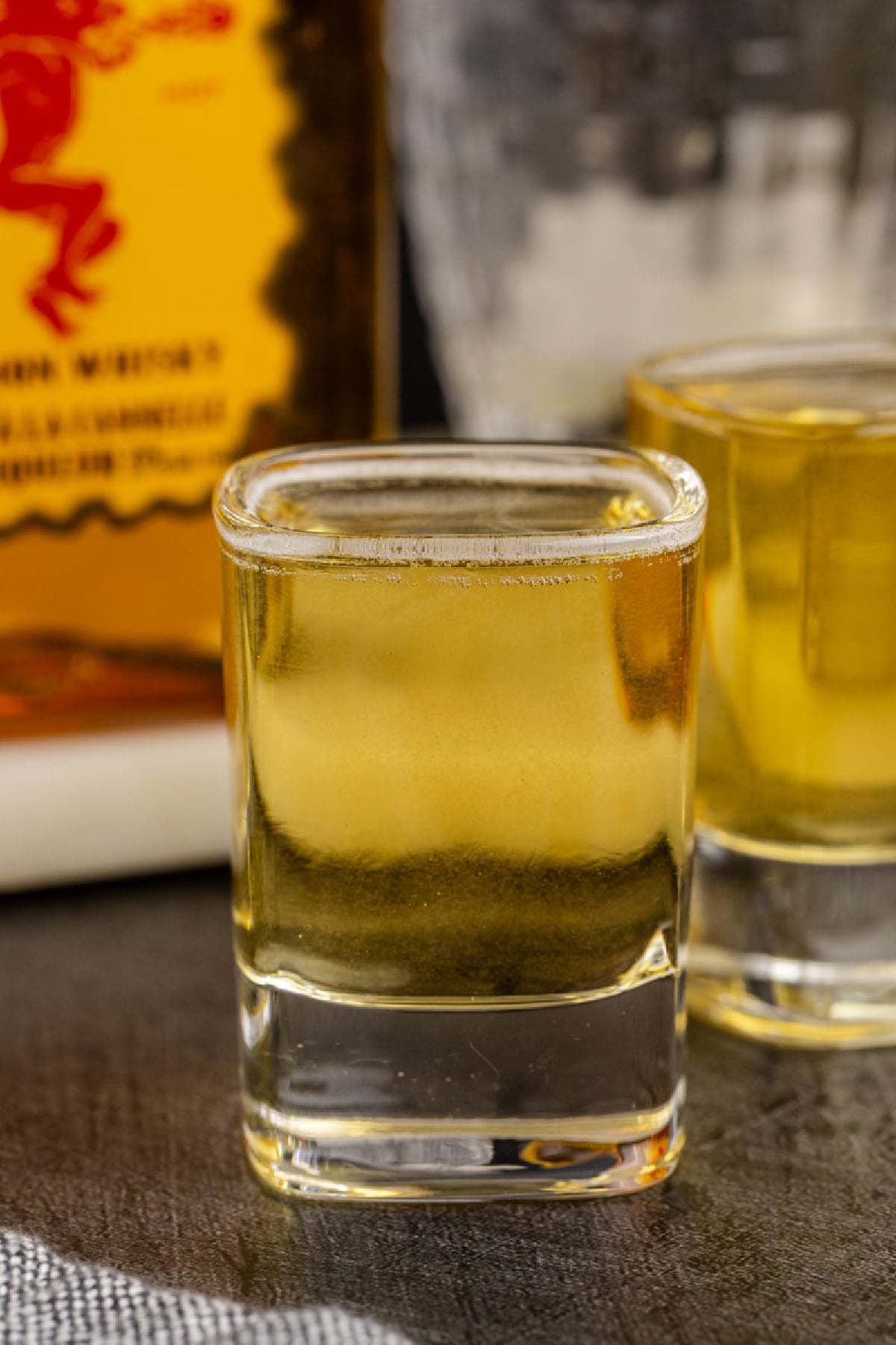 Two shots on a table with a bottle of whiskey in background.