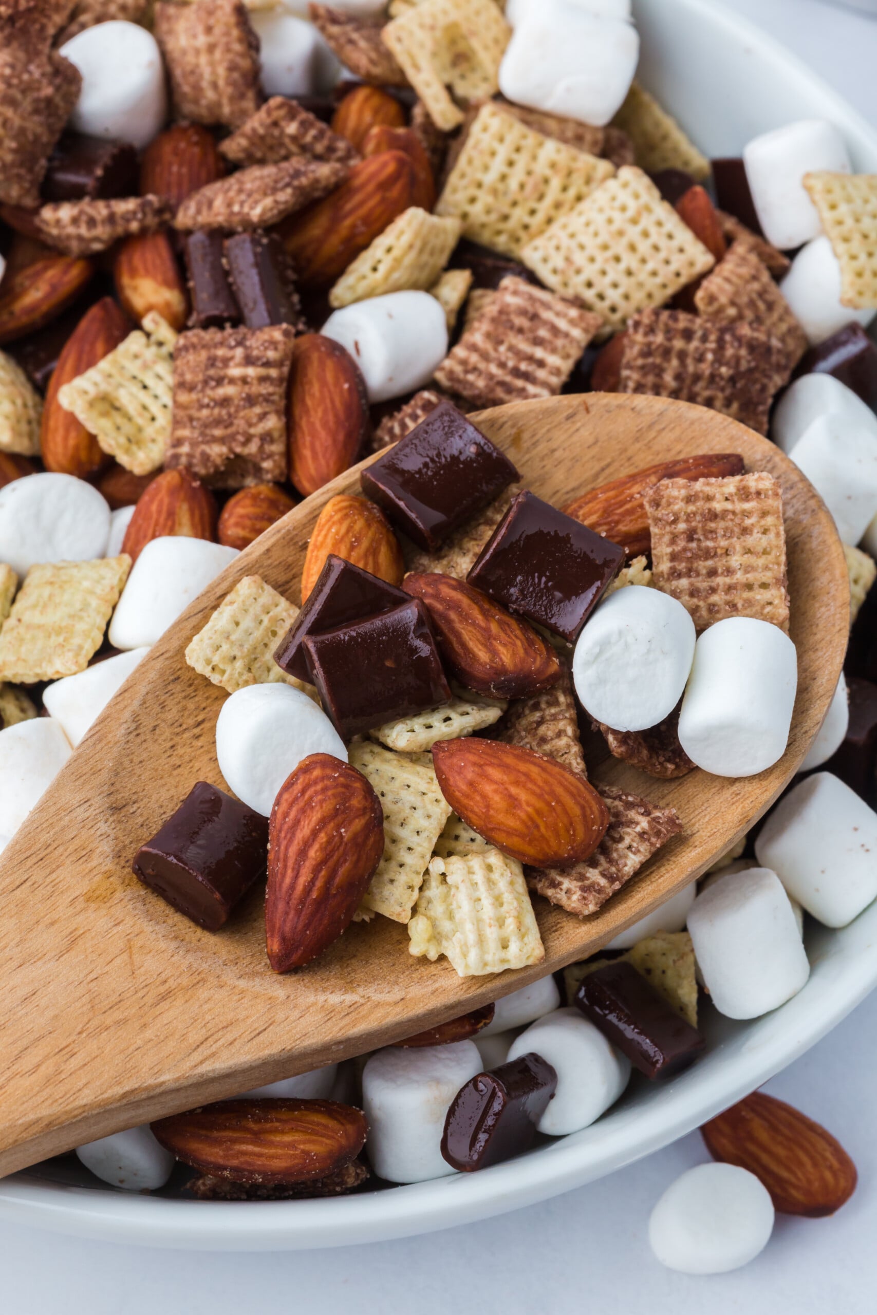 A wooden spoon filled with Trail Mix over a bowl of Chocolate Chex, marshmallows, almonds, and chocolate chunks.