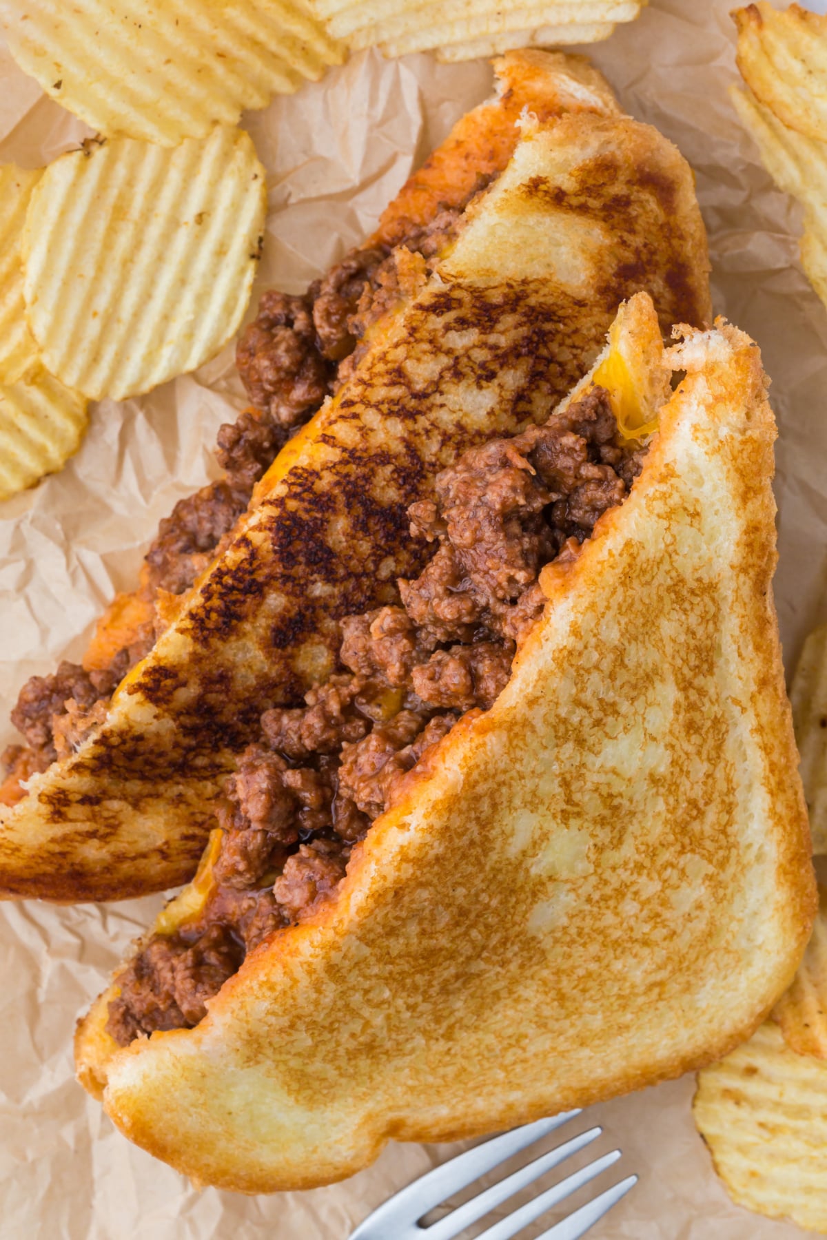 Two sloppy joe grilled cheese halves leaning on one another on parchment paper with chips.