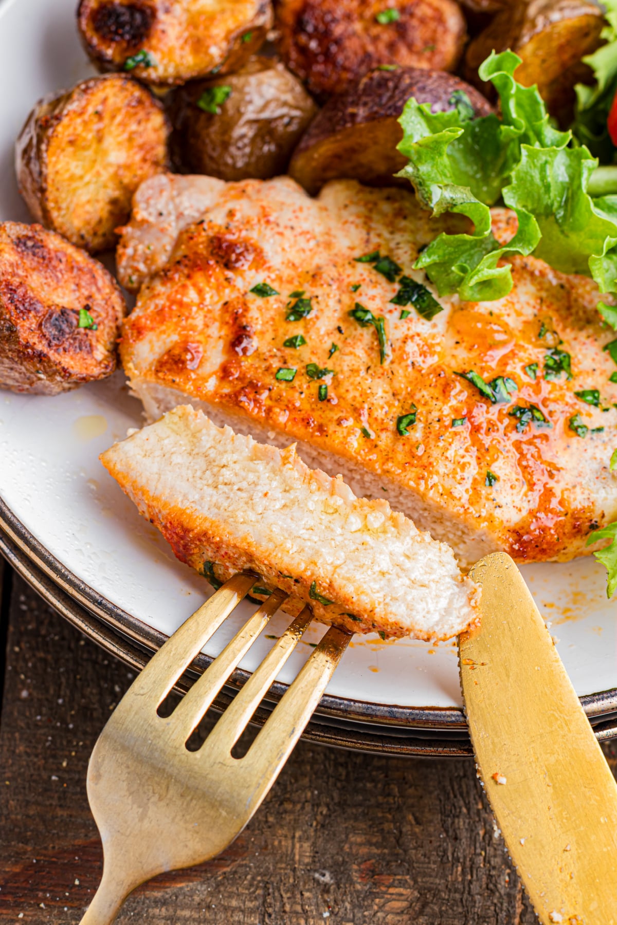 A fork and knife cutting a piece of pork chop with a side of potatoes.