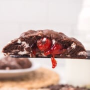 One of the cherry chocolate cookies being held up in the air with a plate of more in the back.