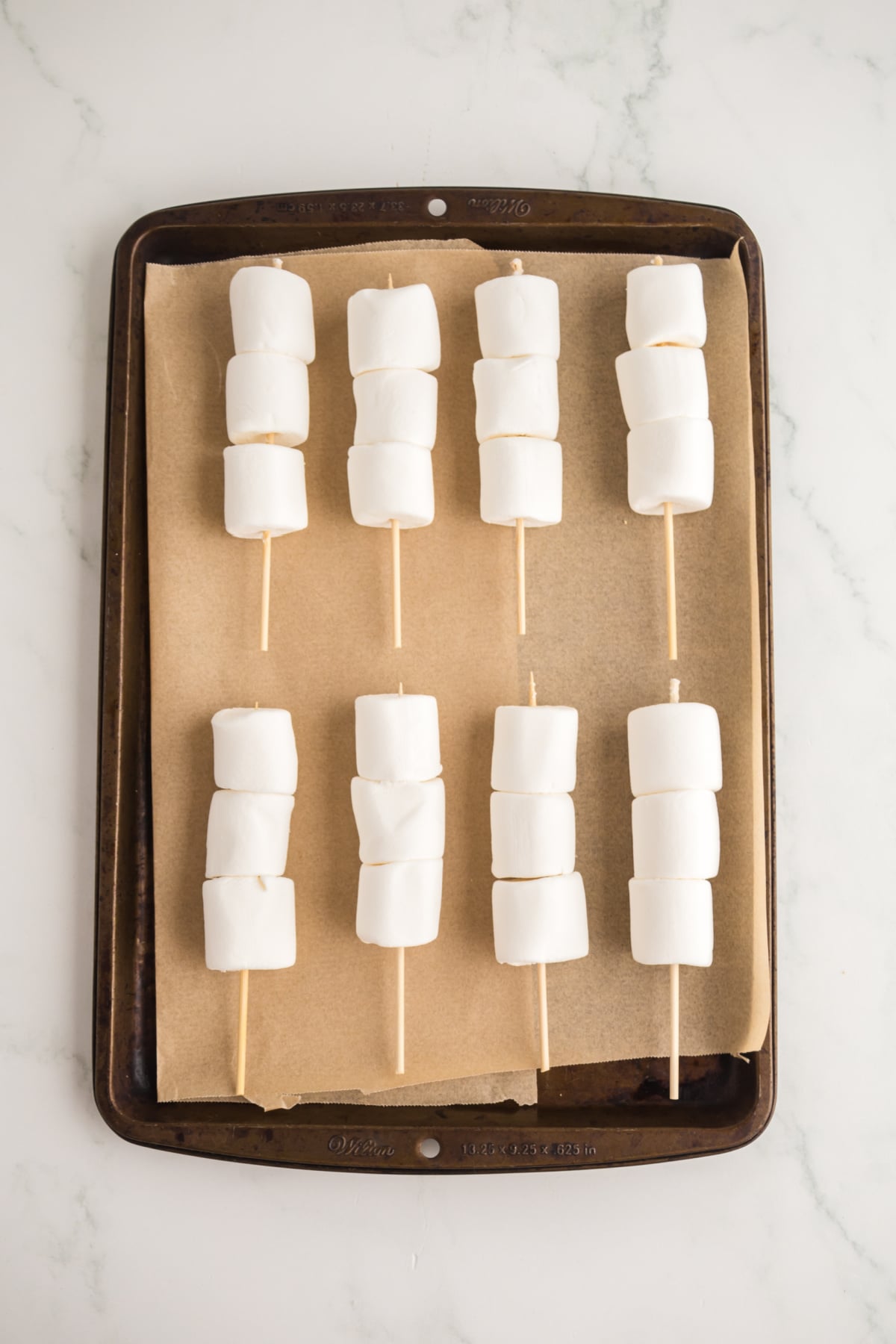 Marshmallows threaded on skewers on a parchment covered rimmed baking sheet.