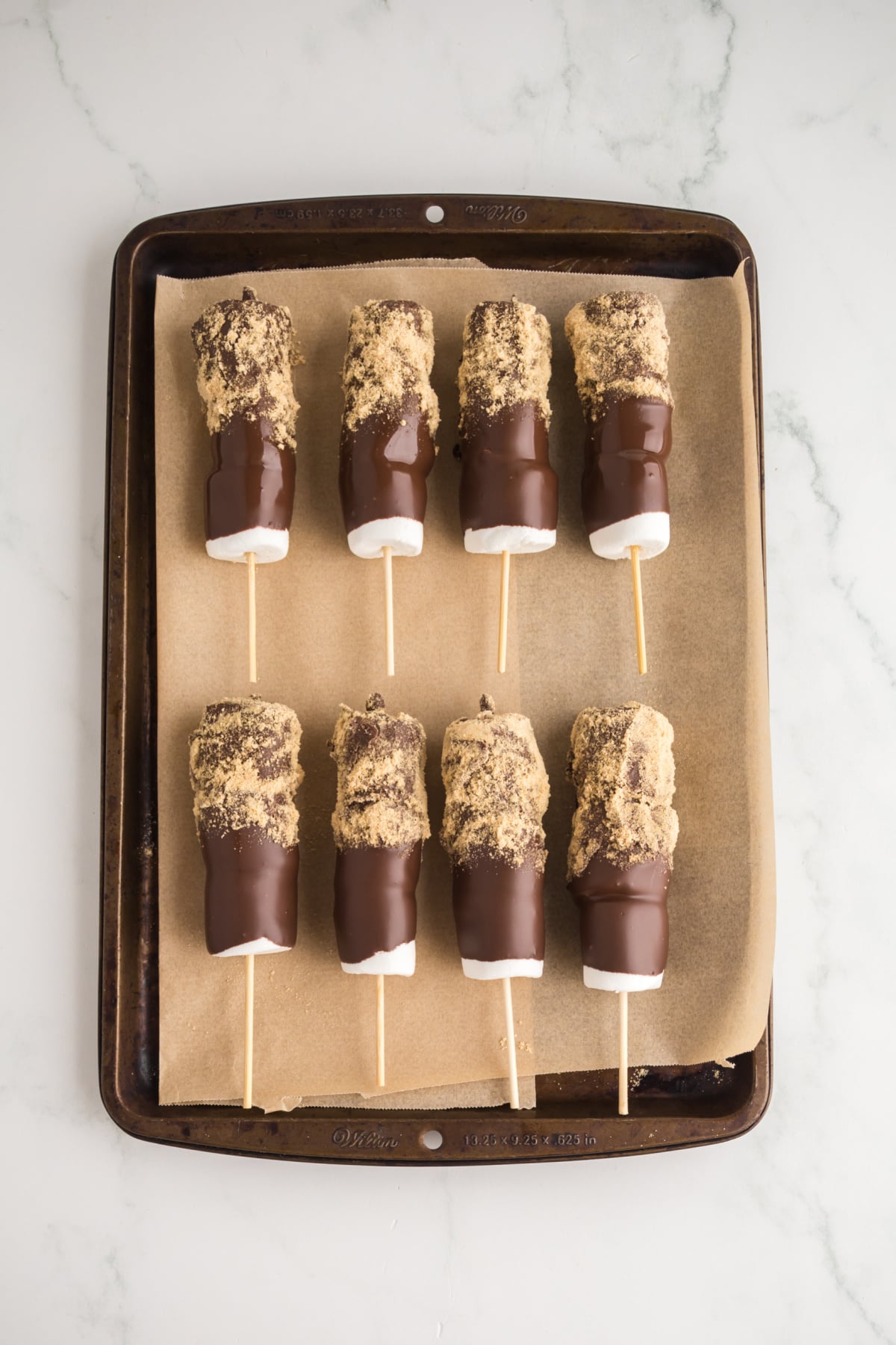 Frozen graham cracker crumb covered marshmallow skewers on parchment covered baking sheet.