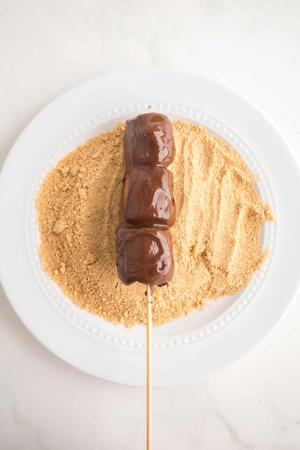Freshly dipped skewer rolling in graham cracker crumbs on a plate.