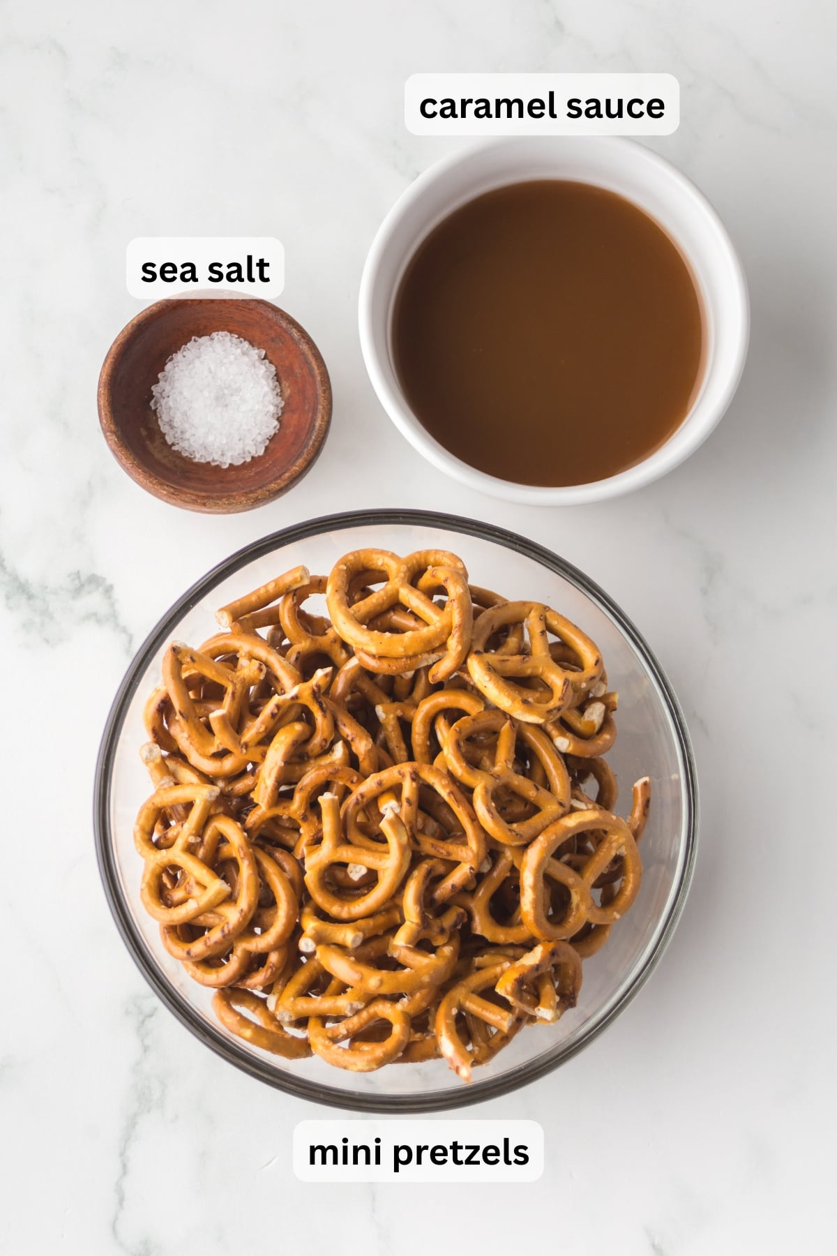 All of the ingredients needed for caramel pretzels including mini pretzels, caramel sauce, and flaky sea salt in bowls on a table.