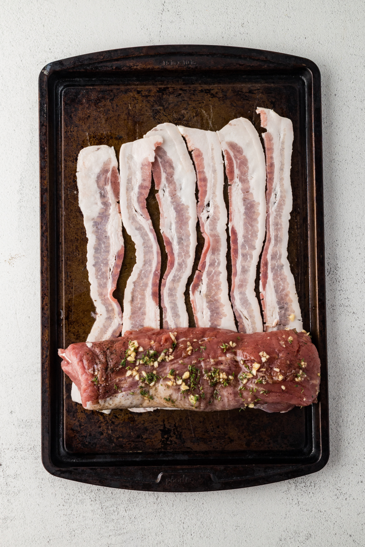 Bacon lined up in a row on cutting board with seasoned pork tenderloin at end ready to roll.