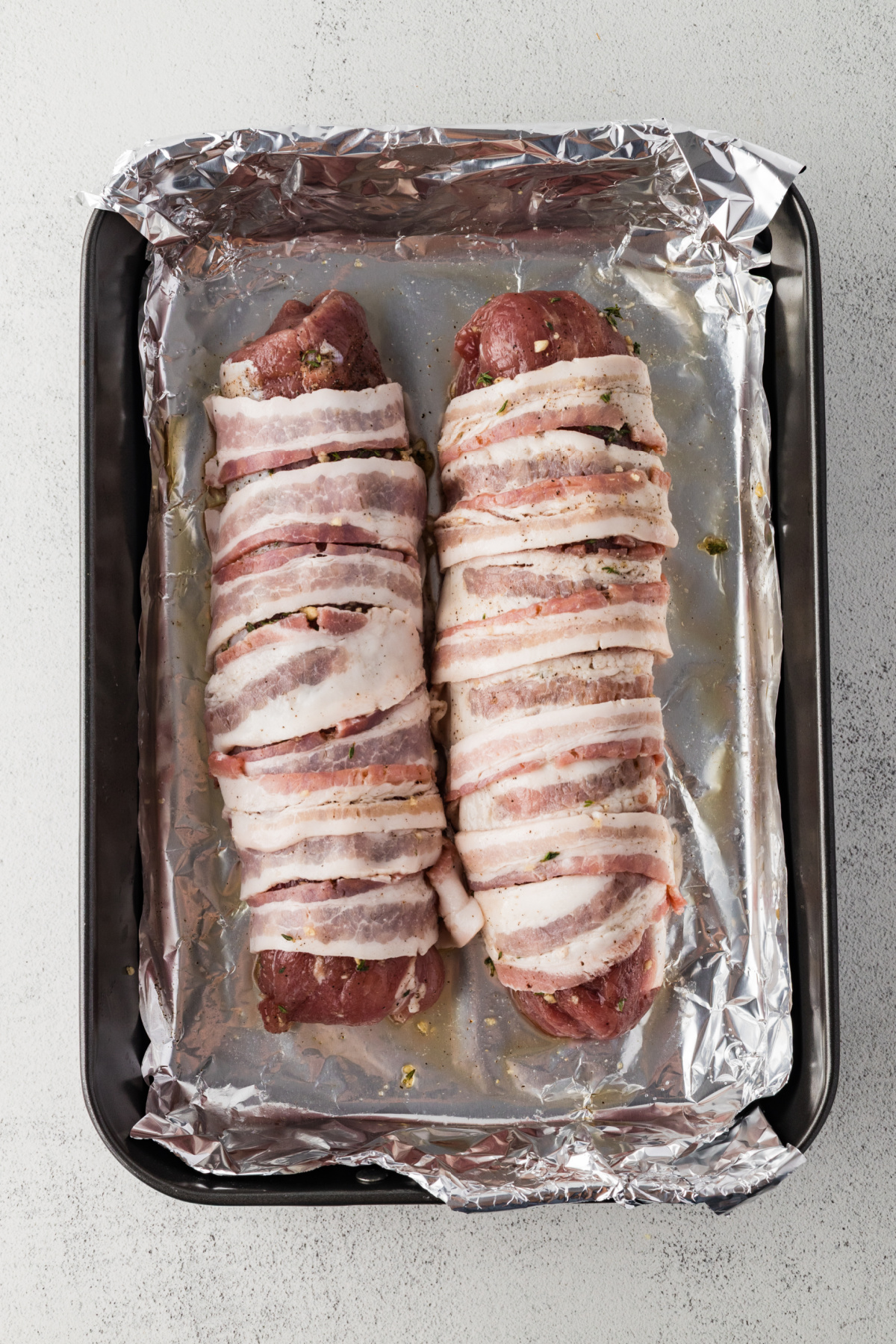 Two tenderloins wrapped in bacon on a foil lined rimmed baking sheet.