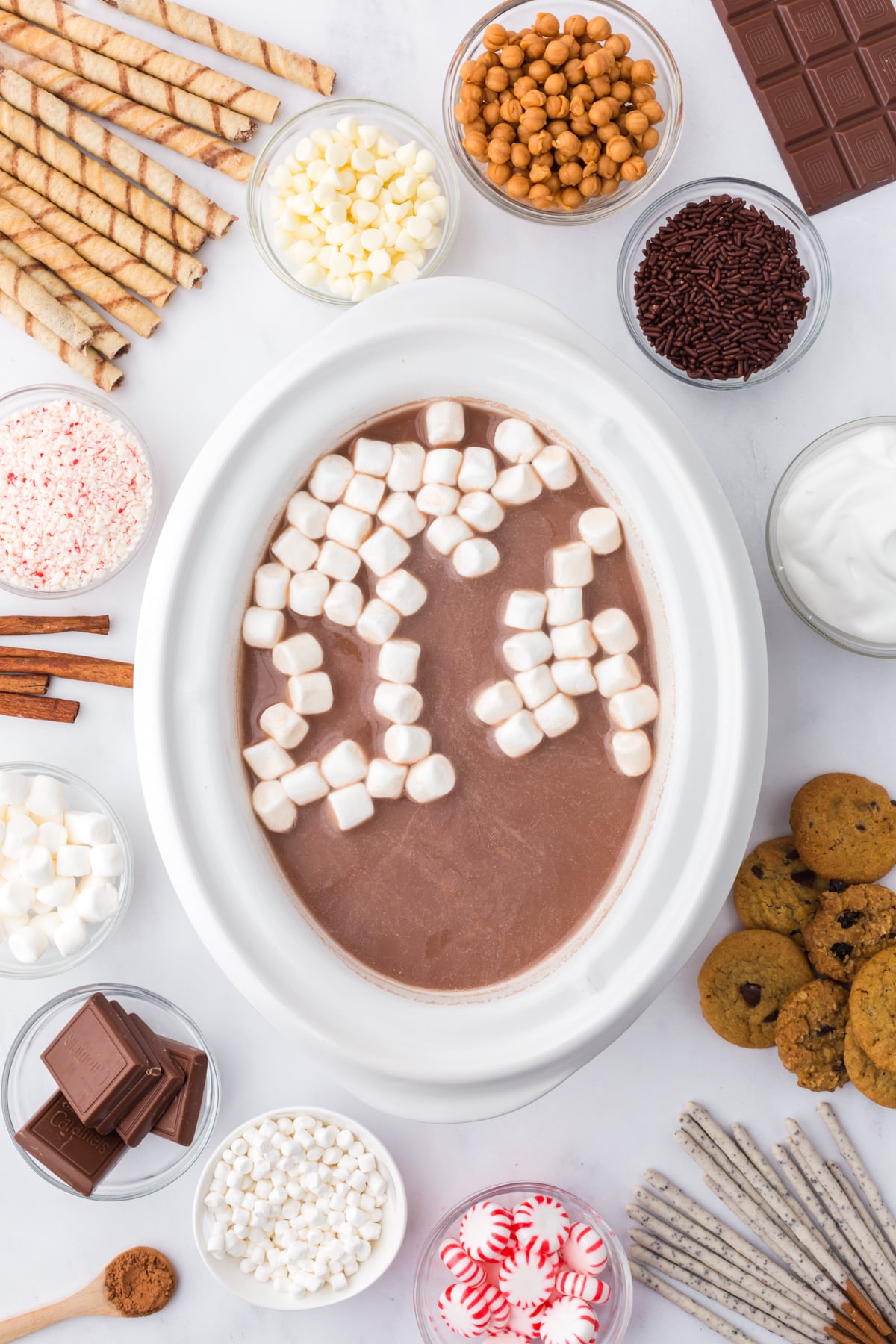 A slow cooker full of hot chocolate surrounded by ingredients needed to create a bar.