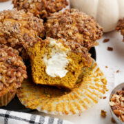 Closeup photo of Pumpkin Spice Muffins with Cream Cheese Filling served in a black plate. One muffin is cut in half revealing the filling. A bowl full of pecans next to the plate.
